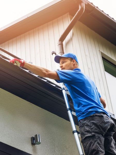 Man in blue shirt cleaning home gutters on a ladder. He wears a blue cap and red gloves, removing debris from the gutter. Home maintenance.