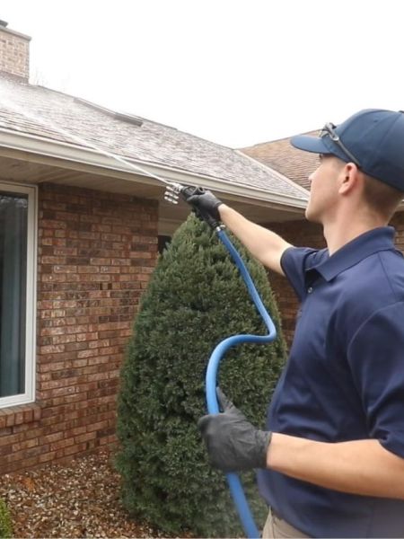 Man cleaning home gutters with a pressure washer, wearing gloves and a cap. The brick house has a visible window and a well-maintained lawn.