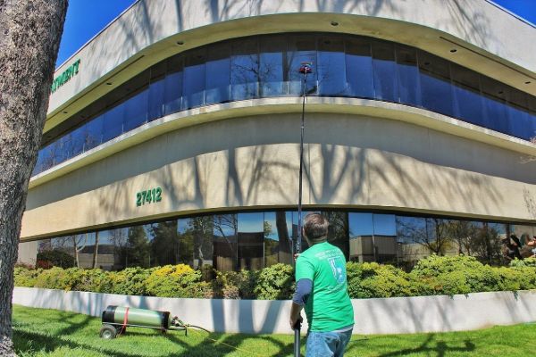 Man using a long pole to perform window cleaning on a large commercial building with reflective windows. The building has the address "27412" visible.